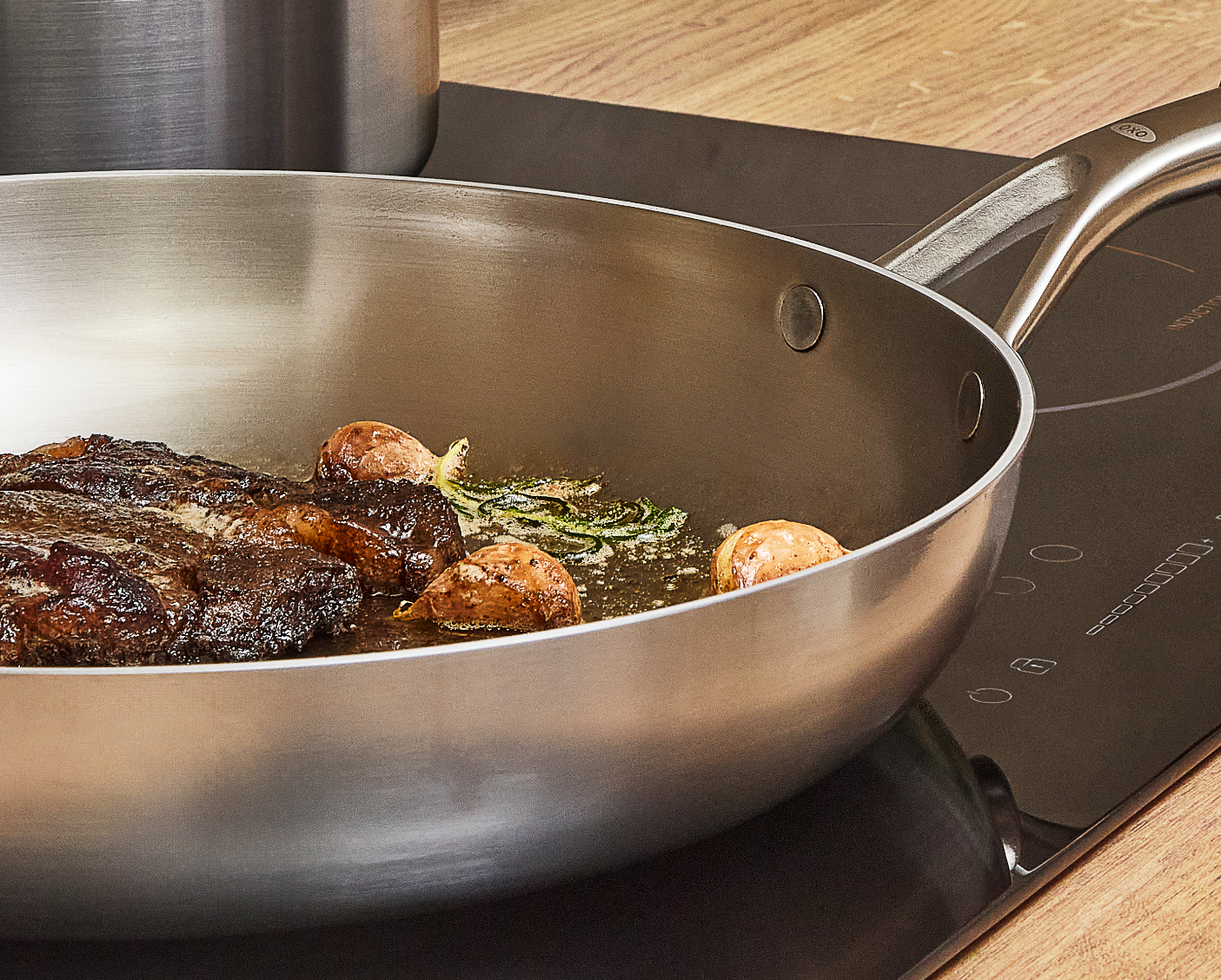 Close‑up of an OXO Mira tri‑ply stainless steel frying pan searing steak with garlic and rosemary on an induction hob.